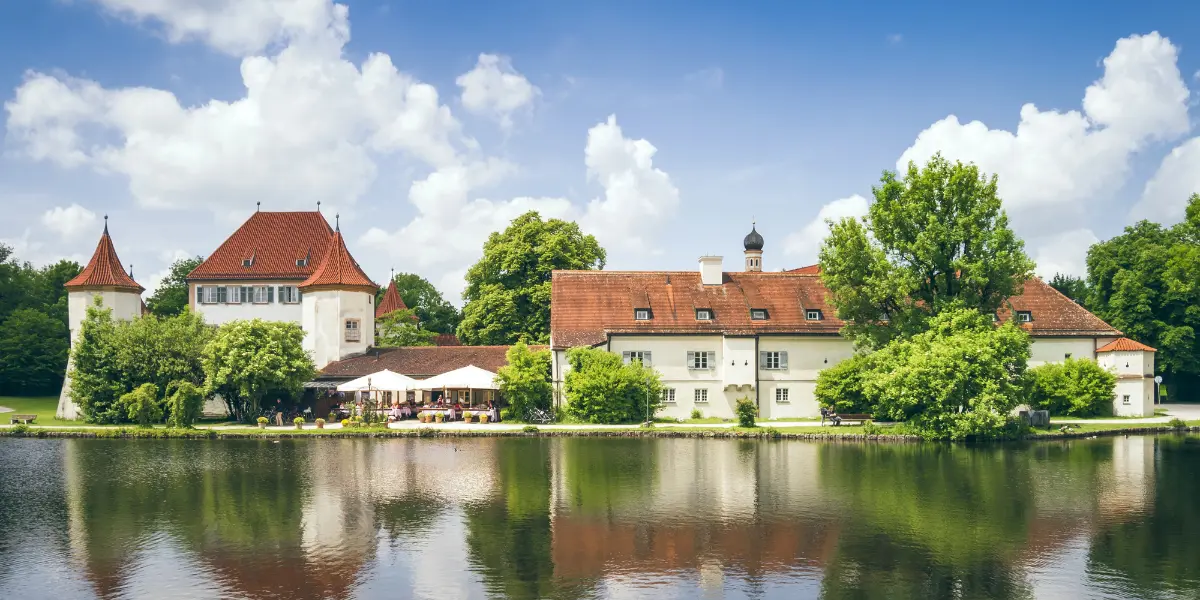 Red-roof Blutenburg Castle under a partly cloudy sky and water in front.