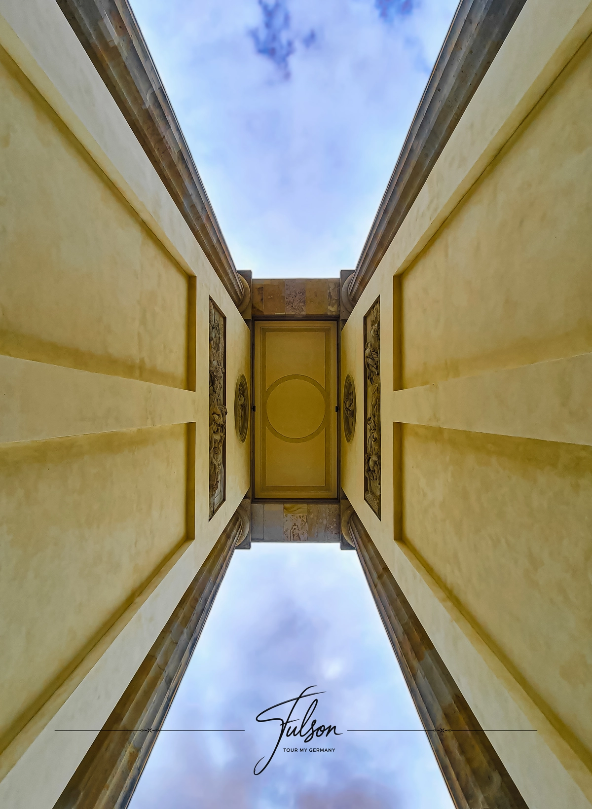 Looking up at a symmetrical architectural Brandenburg Gate in Berlin with detailed panels, flanked by walls, against a partly cloudy sky.