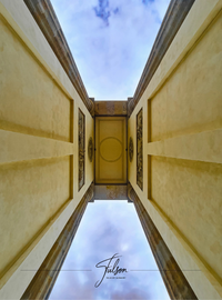 Looking up at a symmetrical architectural Brandenburg Gate in Berlin with detailed panels, flanked by walls, against a partly cloudy sky.