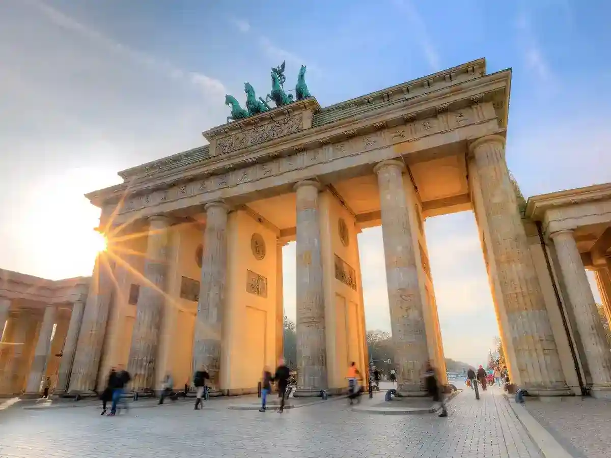 The image shows the Brandenburg Gate in Berlin, Germany, at sunset. Tourists are walking nearby, and the sky is partly cloudy.