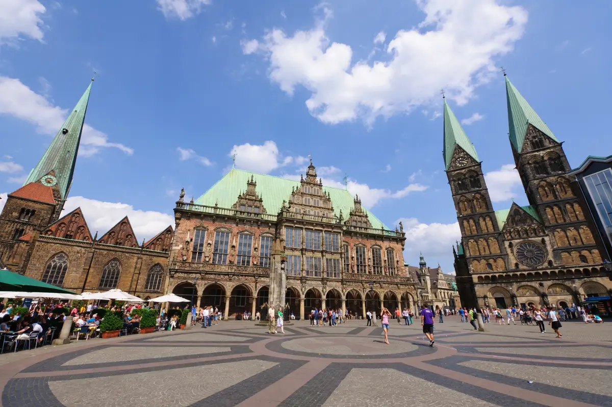 A historic town square with people walking. Prominent buildings feature Gothic architecture, characterized by pointed arches and detailed facades. A clear blue sky is overhead.