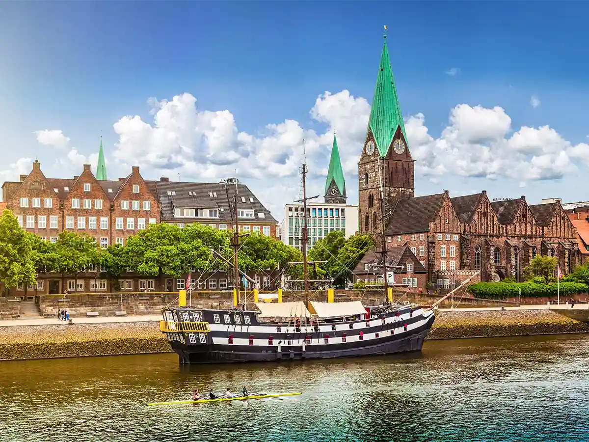 A historic sailing ship is docked on a river in front of old brick buildings and church spires in Bremen; people are rowing in the water under a blue sky with clouds. A historic sailing ship is docked on a river in front of old brick buildings and church spires in Bremen; people are rowing in the water under a blue sky with clouds.
