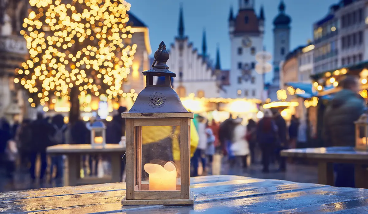 A lantern with a candle on a wooden table at a festive outdoor market is surrounded by people and illuminated by tree and building lights in the evening.