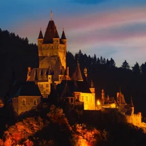 Cochem Castle glowing at twilight, perched above the Moselle River with forested hills silhouetted in the background