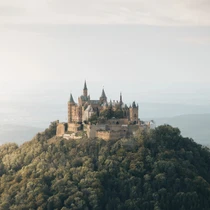 Aerial view of Hohenzollern Castle emerging from forested hills, bathed in soft light under a hazy sky