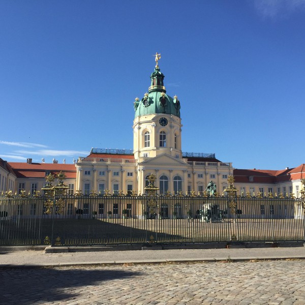 Charlottenburg Palace, Berlin A large historic building with a central dome and clock tower, surrounded by a decorative iron fence, under a clear blue sky.