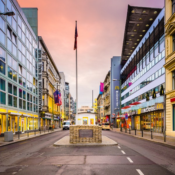 Checkpoint Charlie, Berlin Photo of Checkpoint Charlie in Berlin, Germany, featuring a street lined with buildings, shops, and the museum to the Berlin Wall, with a border guard booth centered and an American flag above.
