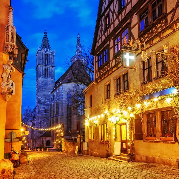 A cobblestone street at sunset lined with historic buildings adorned with lights, a church with pointed towers in the background.