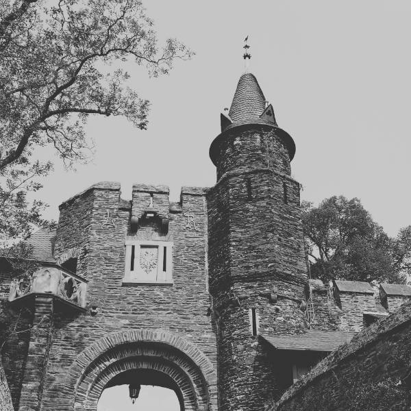 Cochem castle old architecture A black and white photograph showcasing the imposing stone entrance gate of Cochem Castle, featuring a rounded tower with a conical roof and an arched passageway beneath crenellated walls, framed by tree branches.