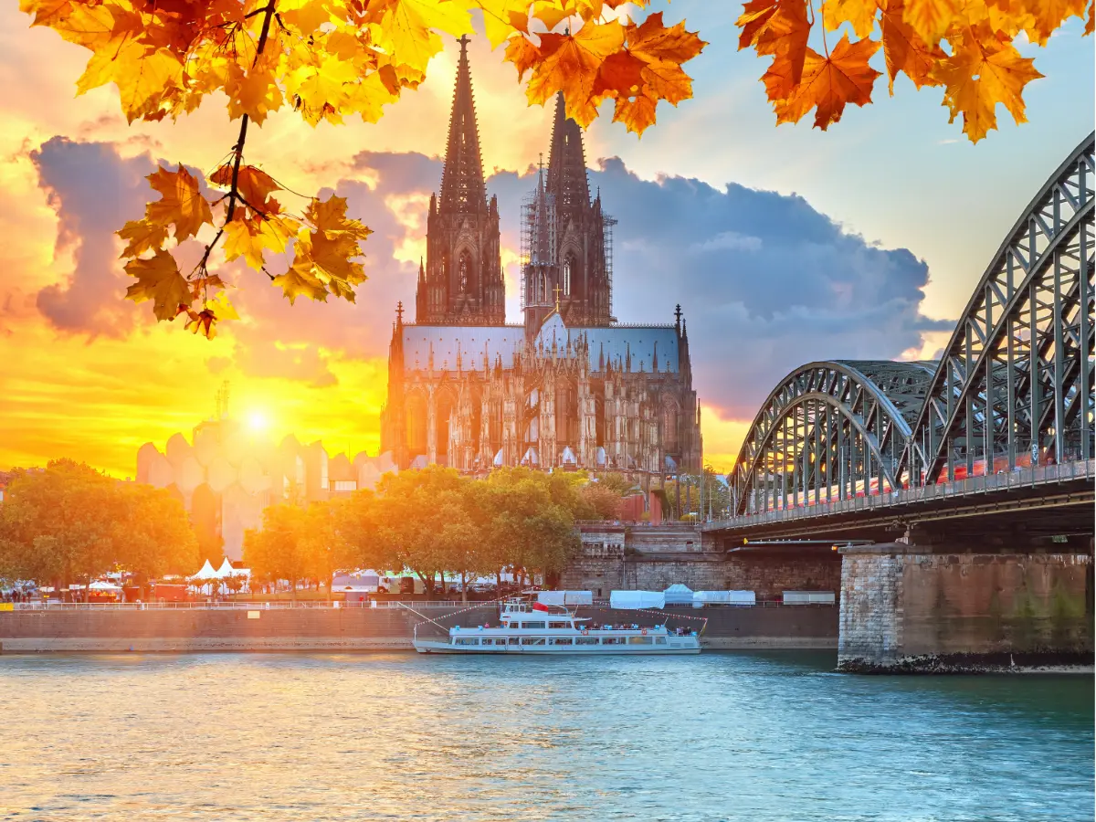 Cologne Cathedral and Hohenzollern Bridge behind autumn-coloured leaves and sunrise.
