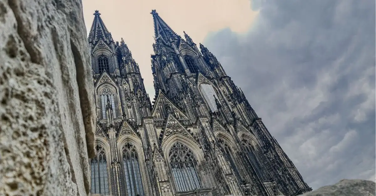 A dramatic upward angle of Cologne Cathedral’s twin spires framed by stormy skies and nearby stone.