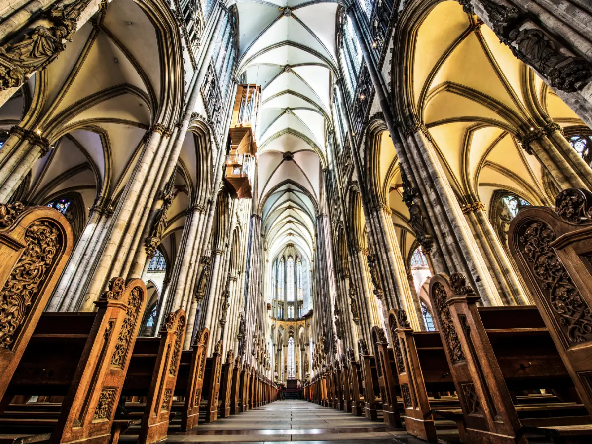 Interior of Cologne Cathedral with towering arched ceilings, ornate columns, and rows of wooden pews leading to a distant altar.