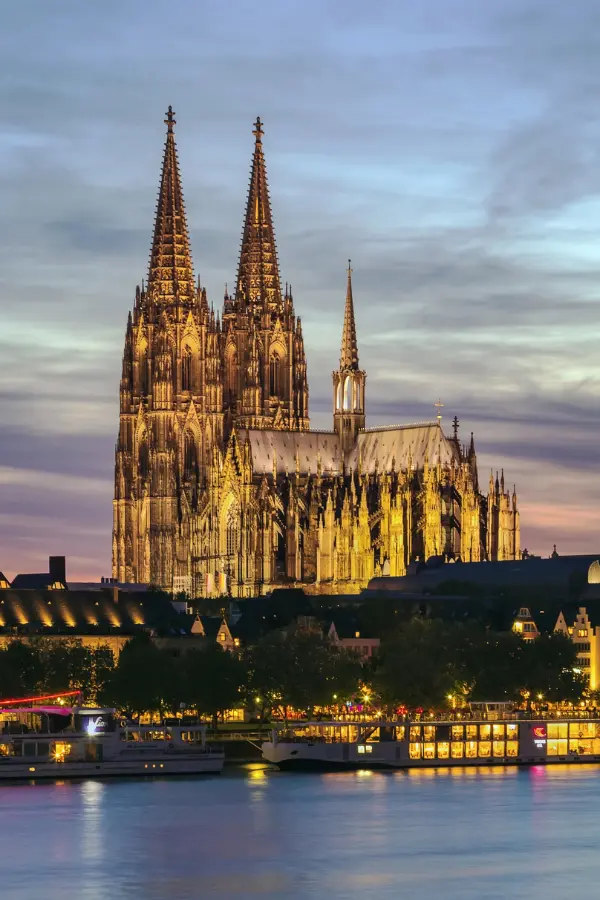 Cologne Cathedral at dusk with illuminated spires, set against a twilight sky. A river with boats is in the foreground.
