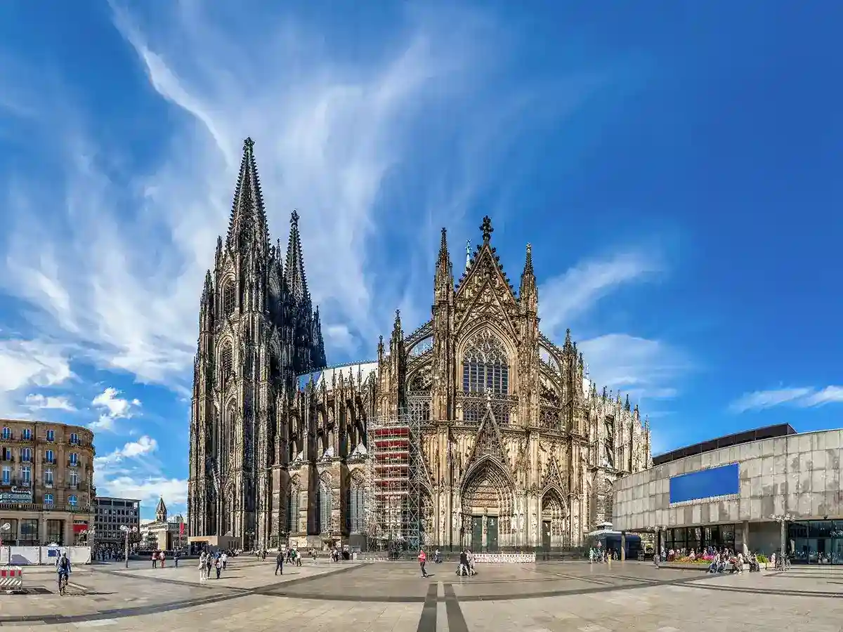 Cologne Cathedral with its twin spires and Gothic architecture, stands in a spacious plaza under a blue sky with scattered clouds. People walk around the square and nearby buildings are visible.