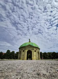 A green-domed pavilion stands under a cloudy sky, surrounded by trees and a gravel path in the foreground.