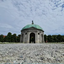 Low-angle view of the Diana Temple in Munich’s Hofgarten, framed by a gravel path and cloudy blue sky