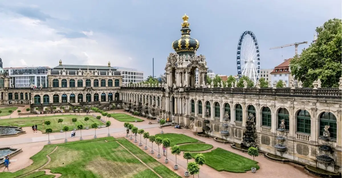 View of the baroque Zwinger Palace in Dresden, Germany, with its ornate crown gate and formal gardens, and a Ferris wheel in the background.