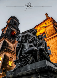 A dark statue depicting a group of figures sits in front of an illuminated historic building with a tall clock tower. The sky is overcast.