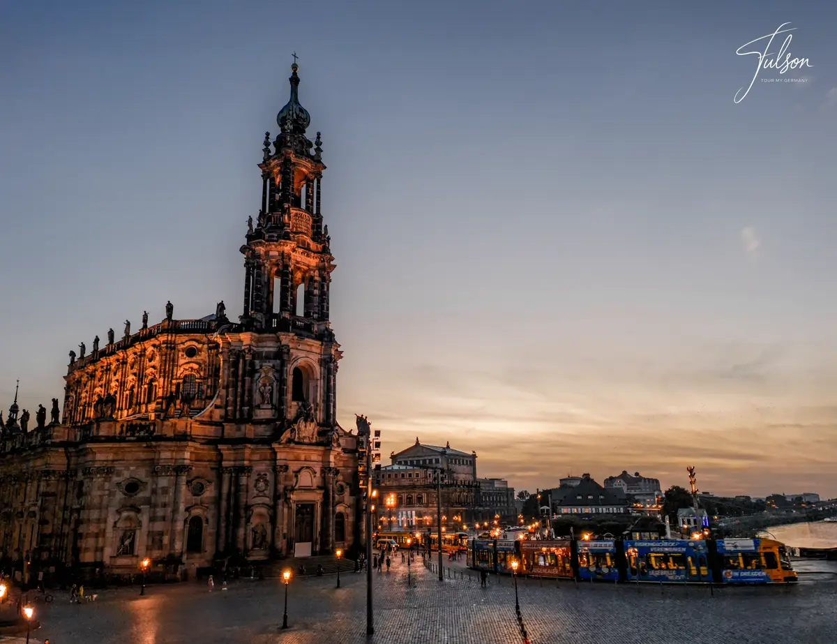 Historic Dresden Cathedral at sunset with streetlights illuminating a plaza, people walking, and a tram passing by under a cloudy sky.