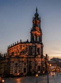 Historic Dresden Cathedral at sunset with streetlights illuminating a plaza, people walking, and a tram passing by under a cloudy sky.
