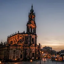 Evening view of Dresden Cathedral lit by warm lights, with a dusky sky and people walking around the square