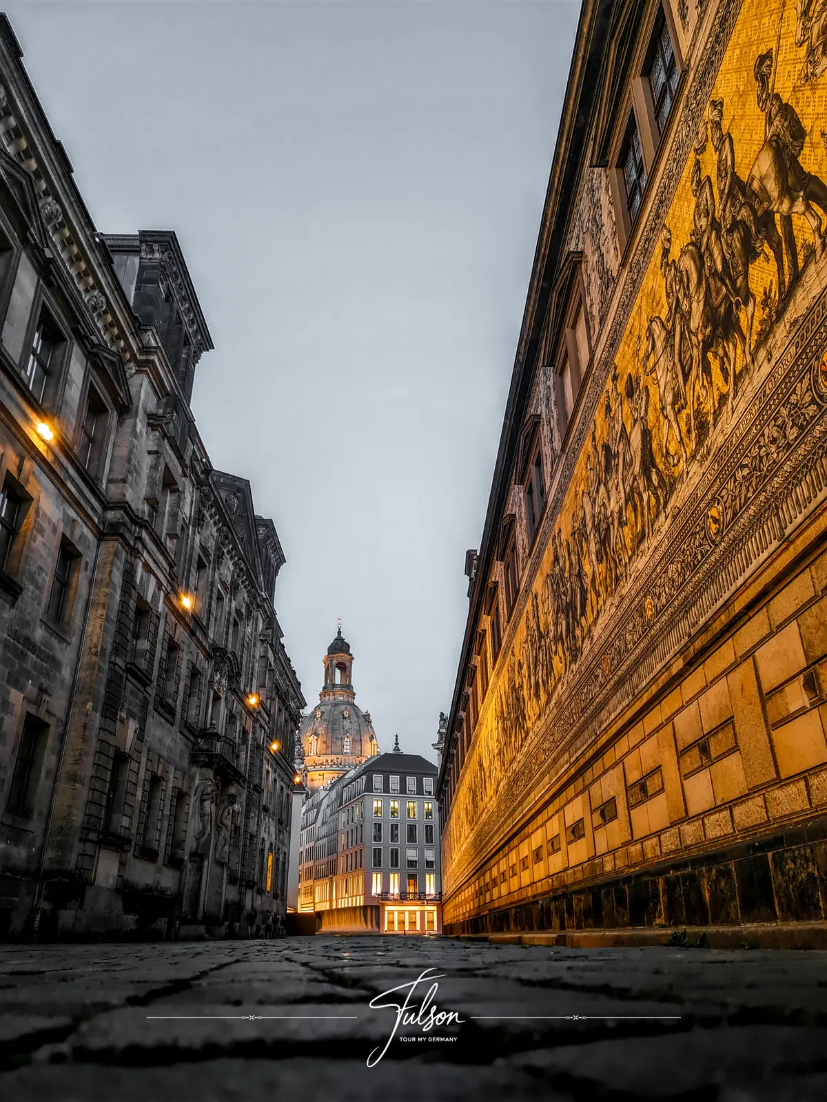 Street view featuring historic buildings and a detailed mural on the right, leading to the distant illuminated dome of Dresden's Frauenkirche under a cloudy sky.