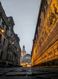 Street view featuring historic buildings and a detailed mural on the right, leading to a distant illuminated dome under a cloudy sky.