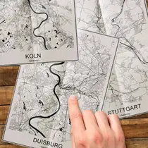 Hand pointing to black-and-white printed maps of German cities including Köln, Duisburg, and Stuttgart.