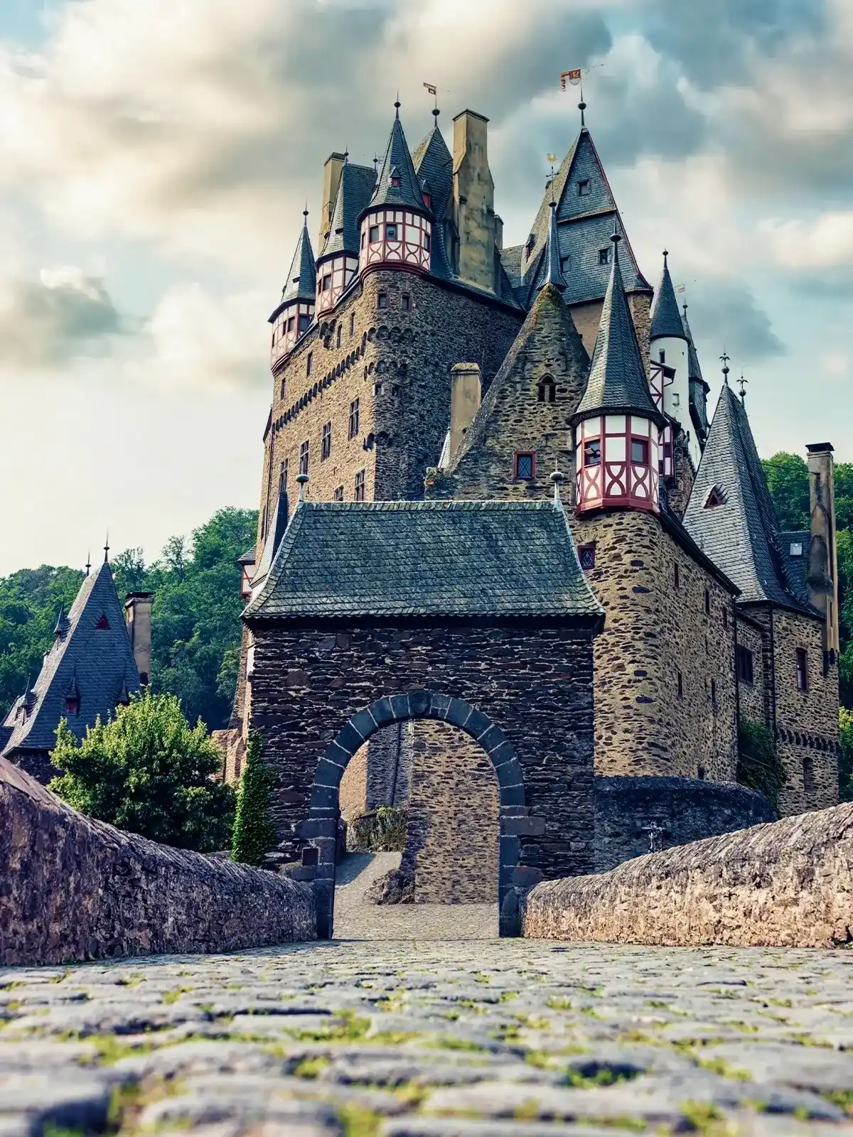 Stone pathway leading to the medieval Eltz Castle with multiple turrets and clouds in the sky.