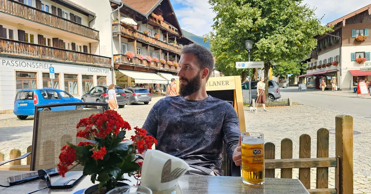 A man enjoys a beer at an outdoor café in Oberammergau, Bavaria, surrounded by Alpine-style buildings and flower boxes.