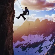 Person rappelling down a steep rock face against a dramatic sunset backdrop and snowy alpine peaks.