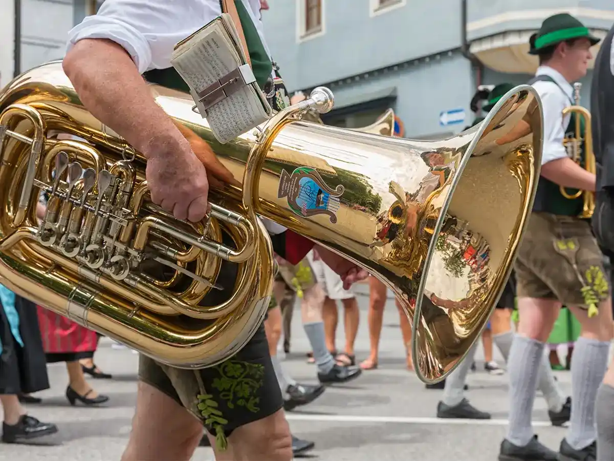 Person playing a tuba in a parade, wearing traditional Bavarian clothing, with onlookers in the background.