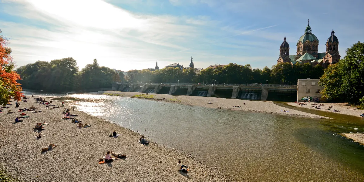 People relaxing along gravel shores of the Isar River with Munich skyline in background