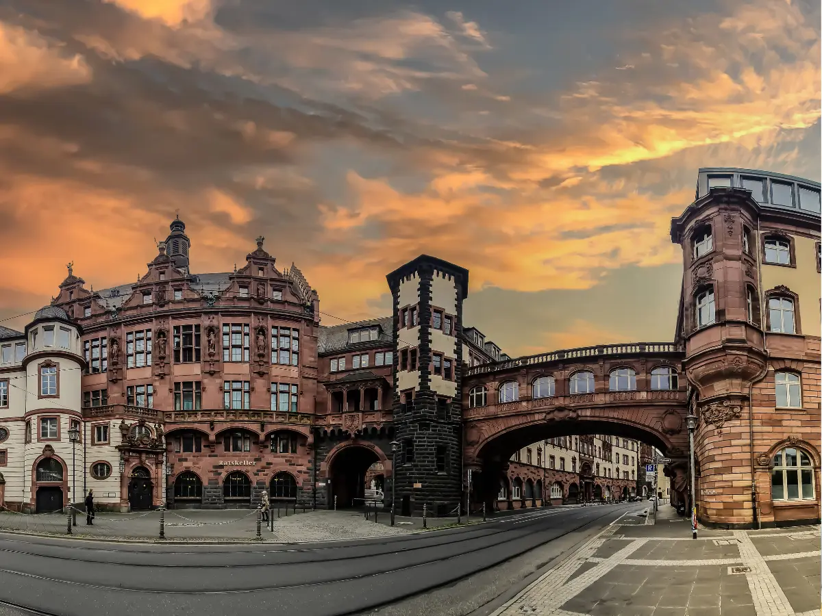 Historic buildings with an arched bridge and intricate architecture stand under a dramatic, colorful sky at sunset in Frankfurt, Germany.