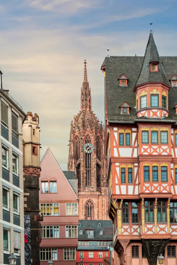 The image shows a skyline with half-timbered buildings in the foreground and a tall Gothic cathedral spire in the background, under a partly cloudy sky in Frankfurt, Germany