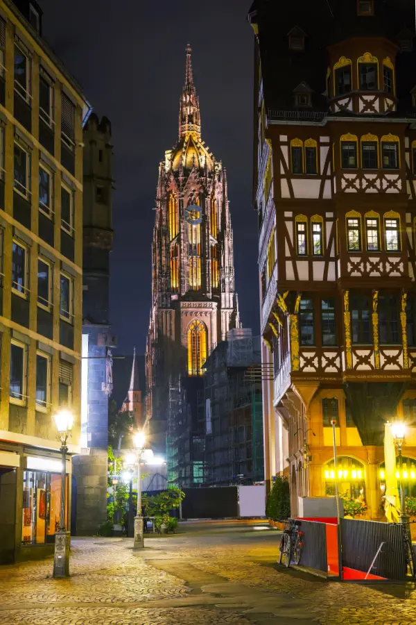 Illuminated Gothic-style cathedral tower at night, framed by half-timbered buildings and streetlamps in a cobblestone square in Frankfurt, Germany.