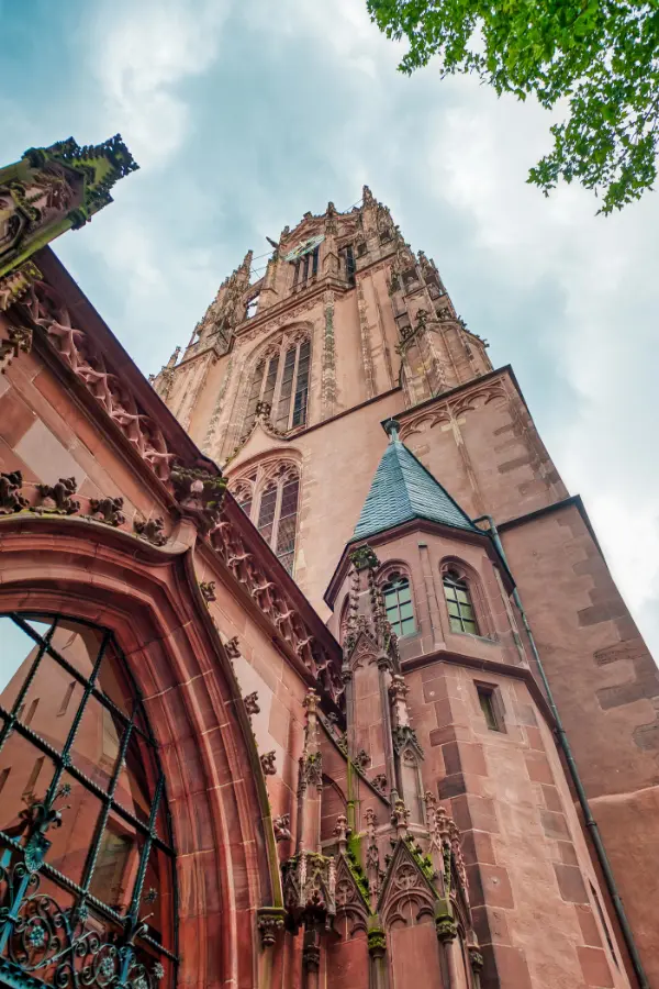 Upward view of the Gothic-style stone Frankfurt Cathedral with intricate detailing, pointed arches, and a tall spire against a cloudy sky. Green leaves frame the top right corner.