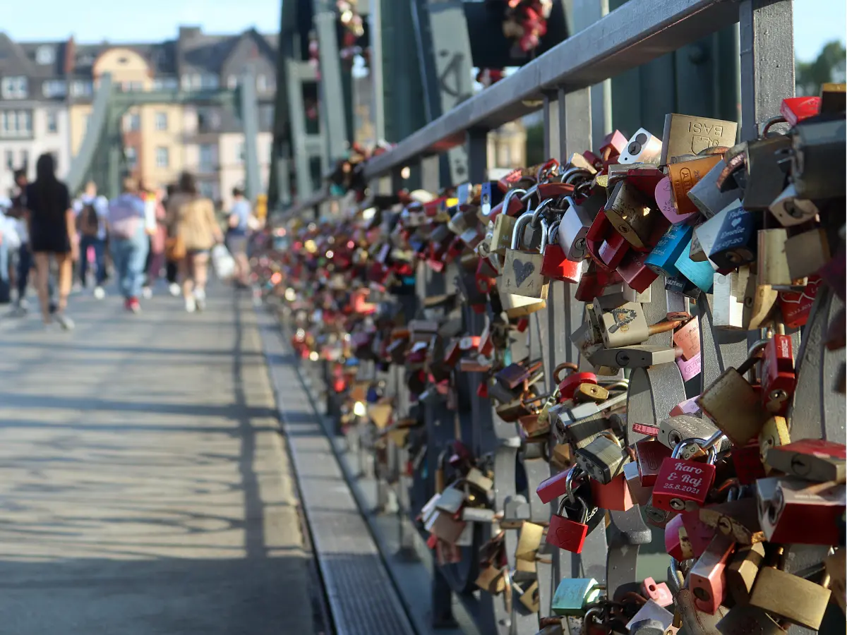 Close-up of numerous colorful padlocks attached to a metal railing on a pedestrian bridge, with people walking in the background.