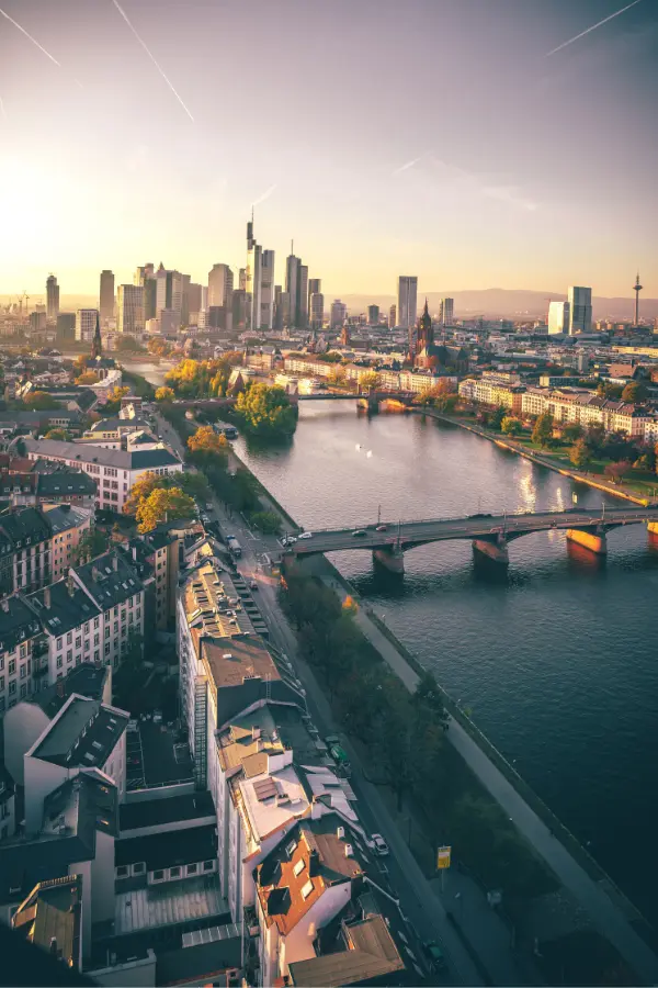 Aerial view of Frankfurt cityscape with skyscrapers, a river, and several bridges at sunset. The scene shows buildings, a calm river, and an illuminated skyline.