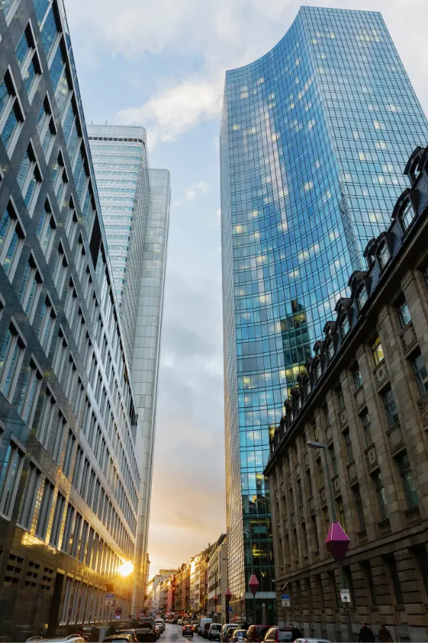 Tall modern skyscrapers in Frankfurt line a city street, reflecting the blue sky and clouds, with the sun setting in the background.