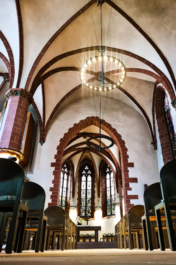 Interior of St. Nicholas Church in Frankfurt with arched ceilings, a circular chandelier, rows of chairs, and stained glass windows at the altar.