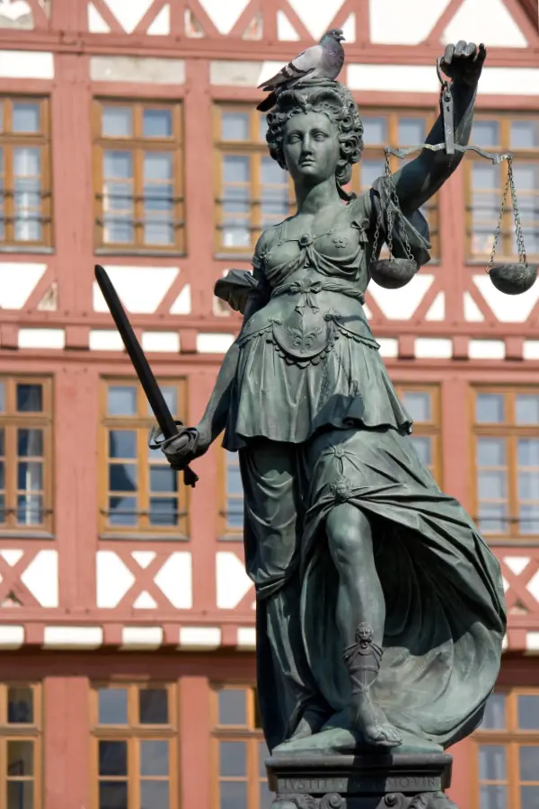 Statue of Lady Justice with scales and sword, a pigeon perched on her head, in front of a half-timbered building.