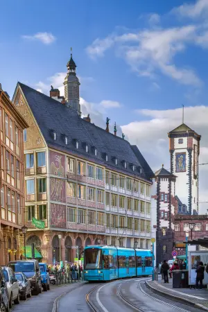 A blue tram on a curved street passes historic buildings adorned with intricate frescoes and a tall clock tower under a blue sky with clouds.
