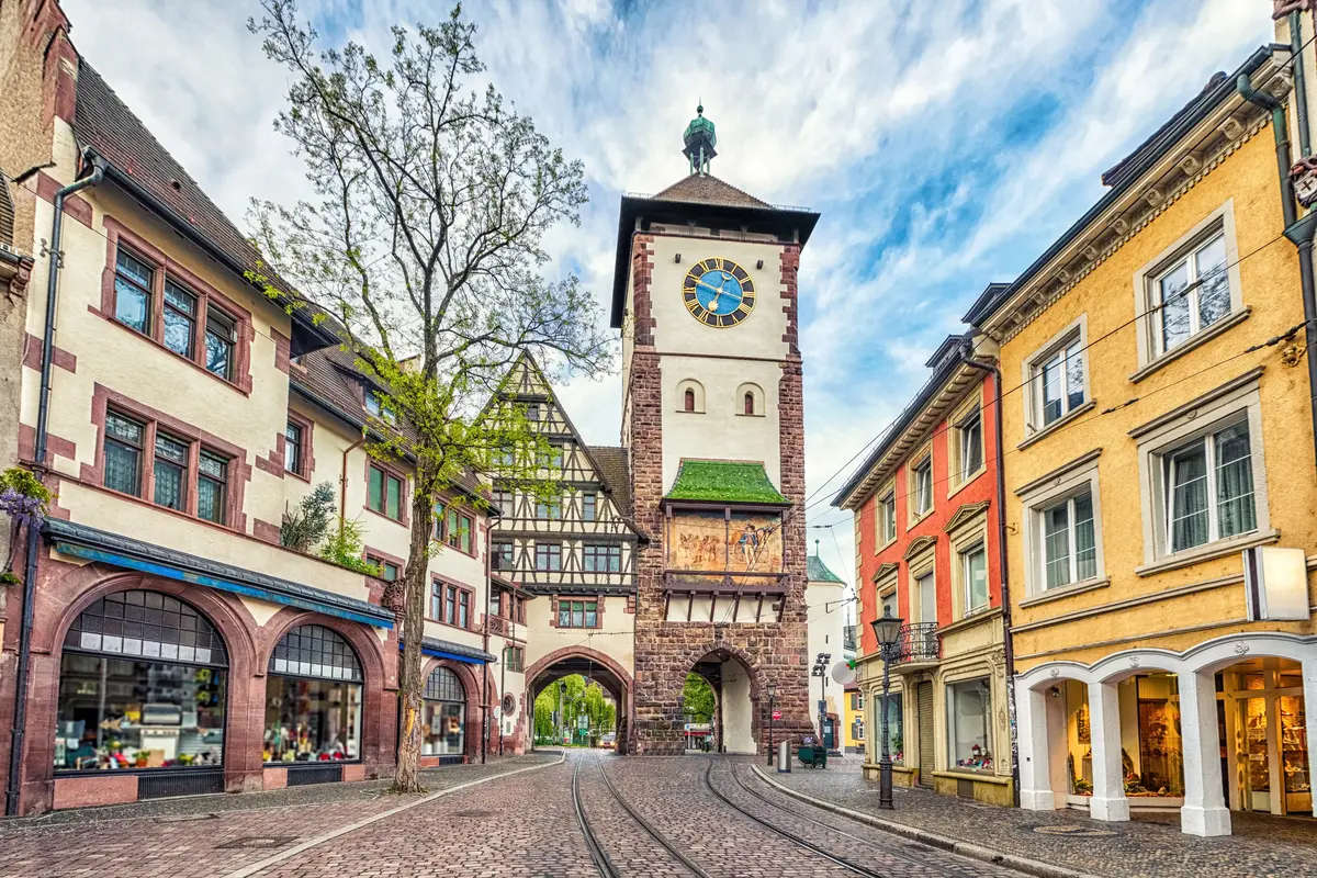 Historic German street in Freiburg with colorful buildings, a cobblestone road, tram tracks, and a prominent clock tower under a blue sky.
