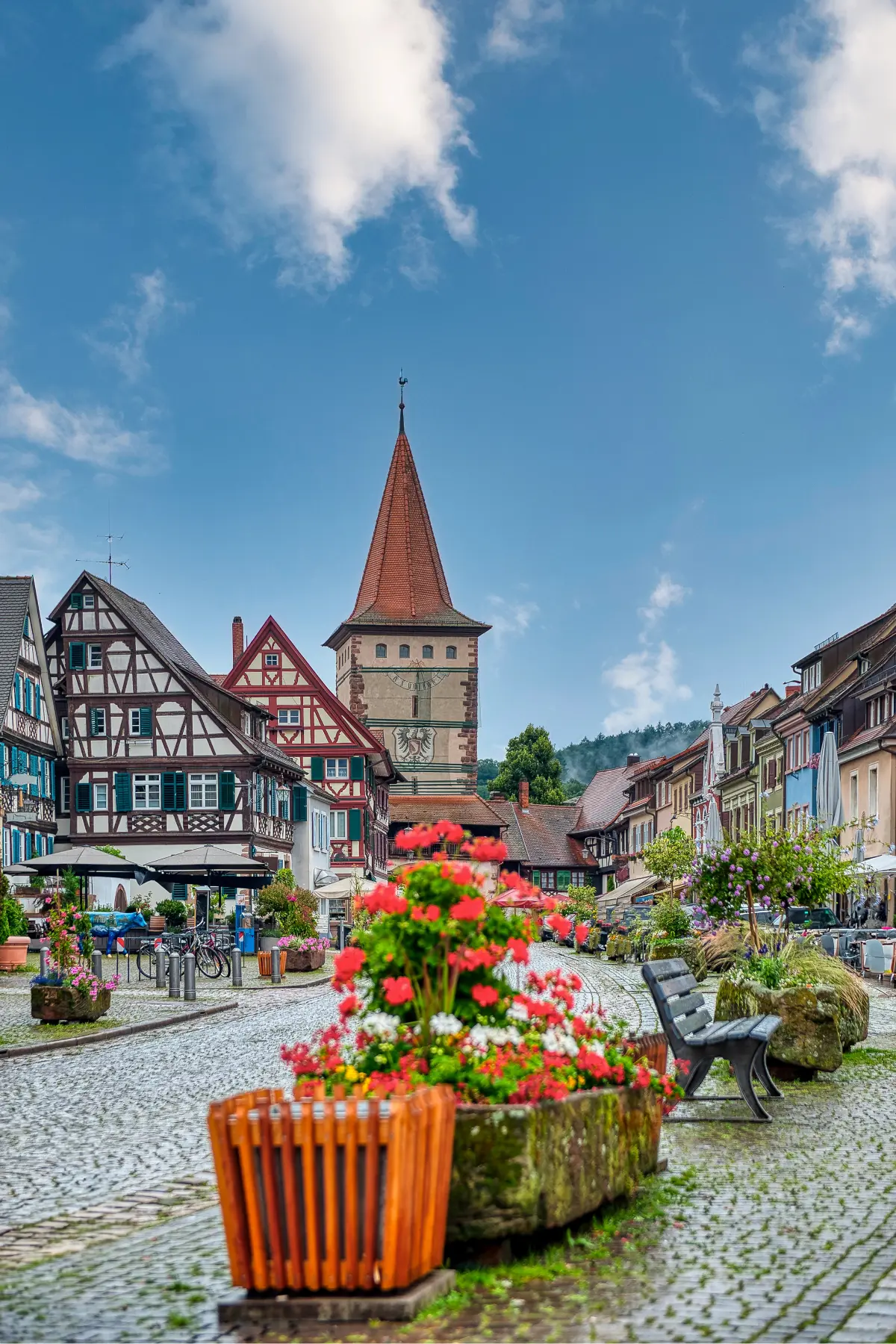 A quaint German village square with cobblestone streets, colorful half-timbered buildings, and a church tower. Bright flowers adorn planters in the foreground.