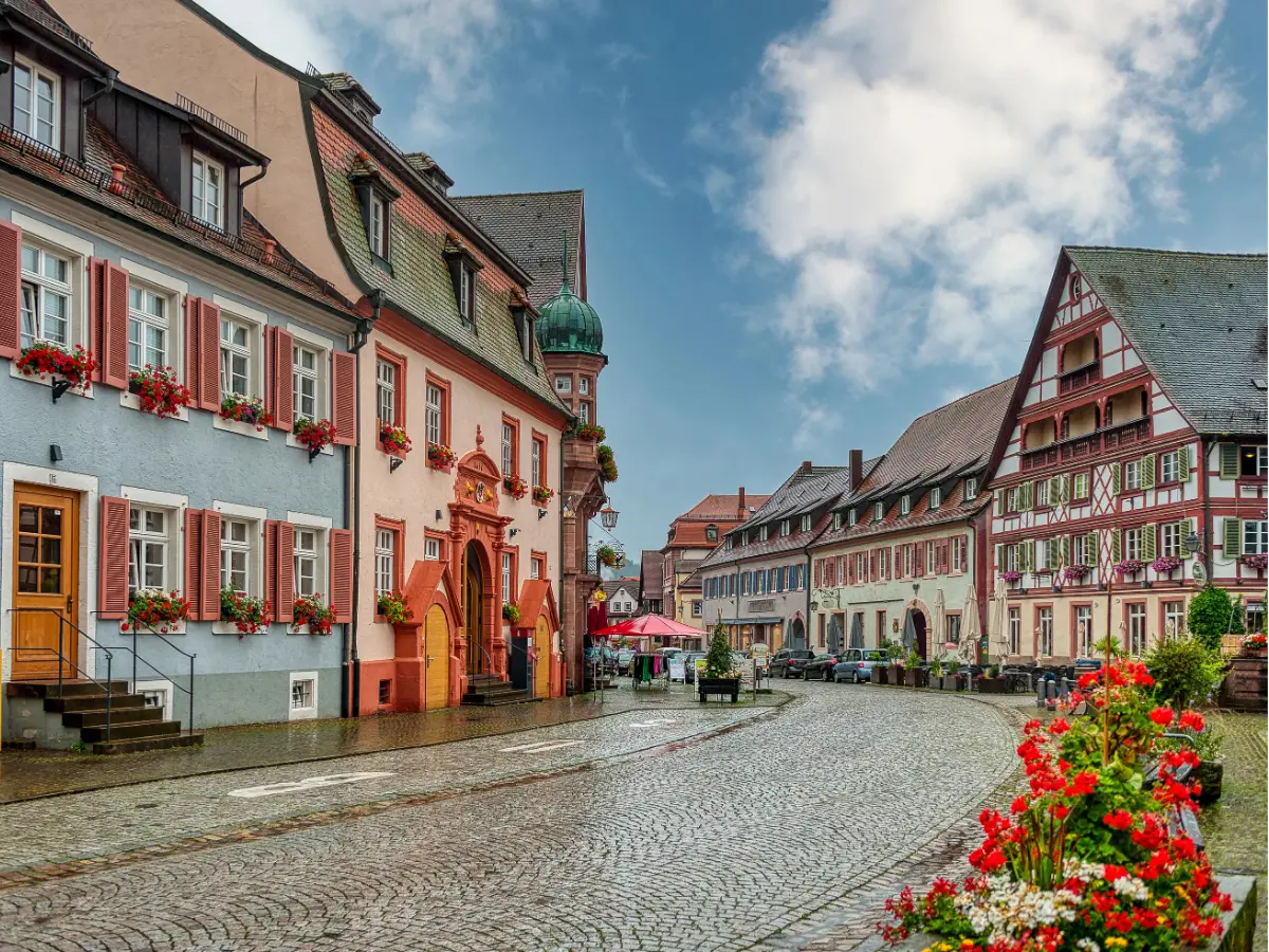 A picturesque European street features colorful historic buildings, cobblestone pavement, and blooming flowers under a partly cloudy sky.