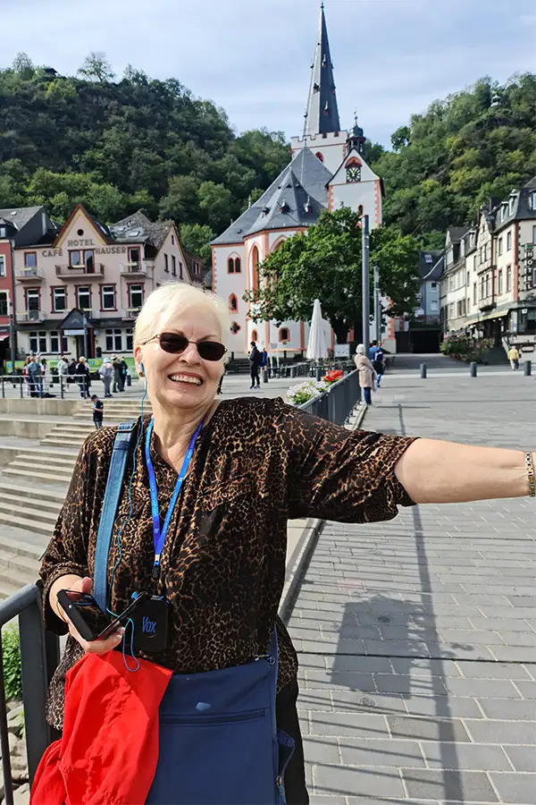 Woman smiling near a riverside promenade with historic buildings and a church in the background on a bright day.