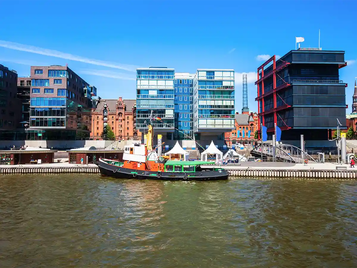 A small boat is docked on a river in front of modern glass buildings and older brick structures under a clear blue sky. A small boat is docked on a river in front of modern glass buildings and older brick structures under a clear blue sky.