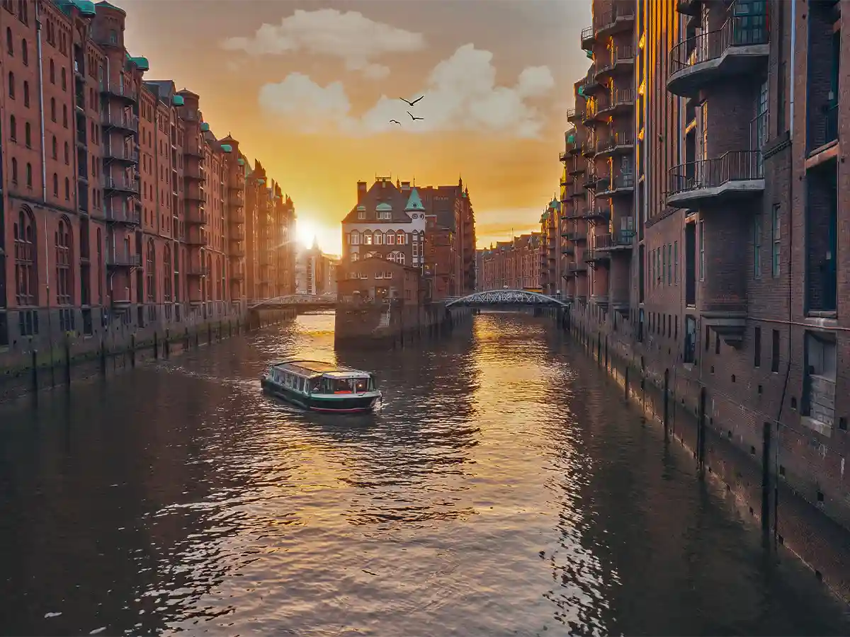 A boat travels along a canal flanked by brick buildings at sunset, with a bridge and distant birds visible under a partly cloudy sky. A boat travels along a canal flanked by brick buildings at sunset, with a bridge and distant birds visible under a partly cloudy sky.