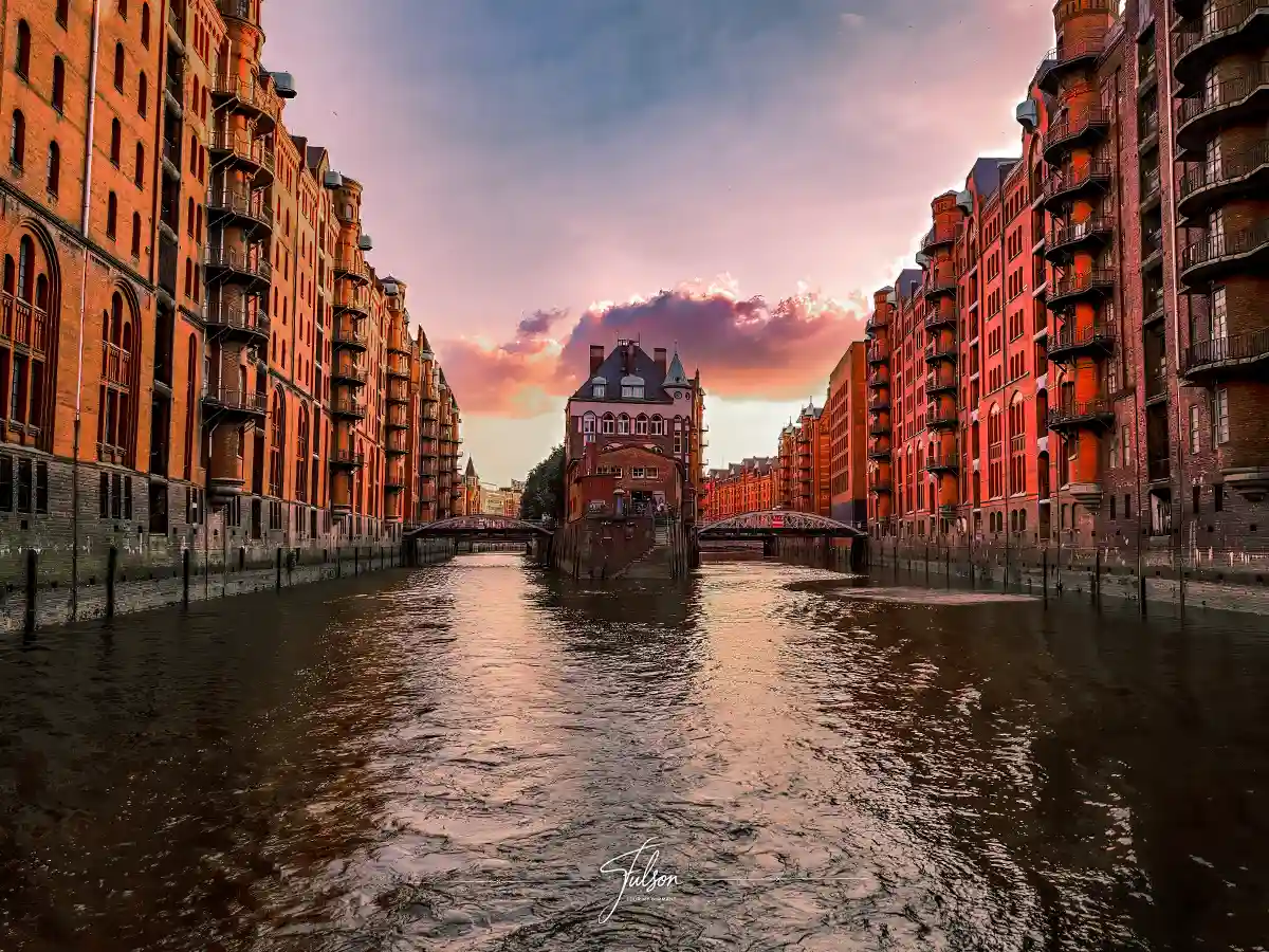 A red-brick building stands at the center of a canal flanked by similar buildings at sunset, with reflections in the water and a cloudy sky overhead. A red-brick building stands at the center of a canal flanked by similar buildings at sunset, with reflections in the water and a cloudy sky overhead.